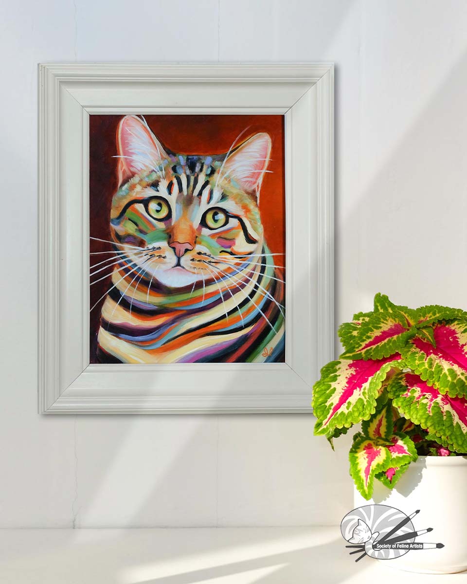 Framed colorful tabby cat painting on a white wall with a plant below.